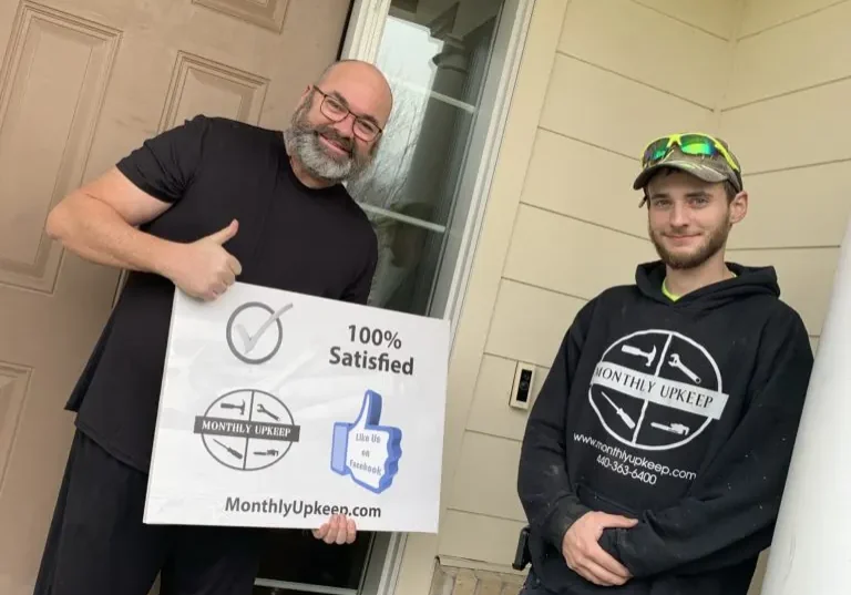 Two men holding a 100% Satisfied Monthly Upkeep sign at a front door, one in a Monthly Upkeep hoodie